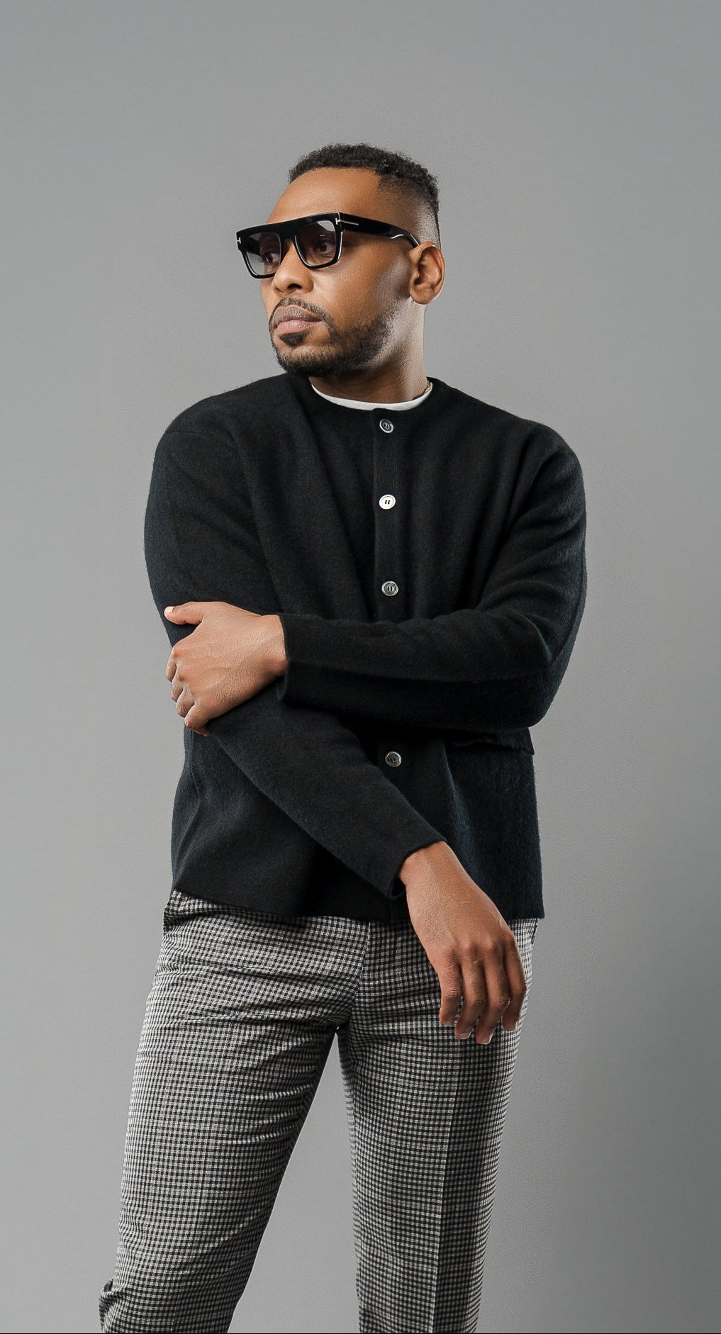 Man wearing a black cardigan, black shirt, gray patterned pants, and black shoes on a gray background